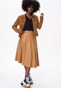 Jupe en cuir beige avec un design évasé, associée à une veste en velours côtelé marron et des baskets noires avec une semelle blanche et orange.