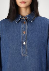 Denimskjorte med en strukturert krage, fem bronseknapper og en svak blek blåfarge. Myk tekstur med synlige sømdetaljer.