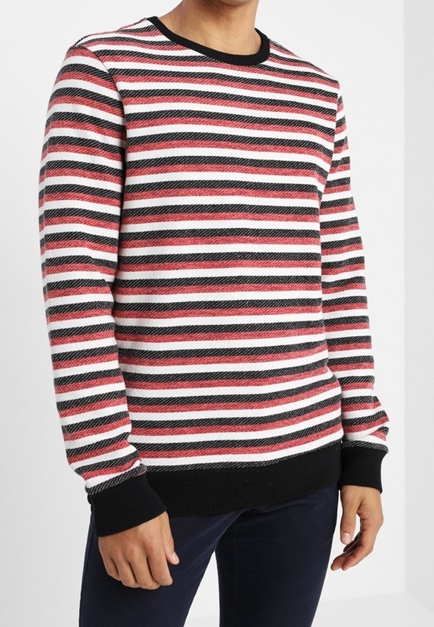 Homme portant un pull à manches longues avec des rayures horizontales rouges, blanches et noires, et des bordures noires aux poignets et à l'ourlet.