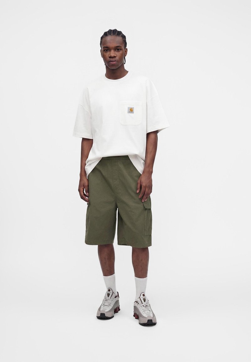 Junger Mann mit geflochtenem Haar, trägt ein weißes oversized T-Shirt, olivgrüne Cargo-Shorts, weiße Socken und graue Sneakers, steht vor einem weißen Hintergrund.