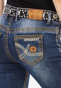 Blaue Jeans aus Denim mit orangefarbenen Nähten, die ein einzigartiges Rücktaschendesign aufweisen, sowie einen schwarzen Gürtel mit silbernen Nieten und einem Lederetikett.
