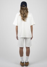 Femme aux longs cheveux ondulés, portant un t-shirt blanc oversize et un short, des chaussettes blanches, des chaussures marron et une casquette de baseball bleu foncé, debout de dos.