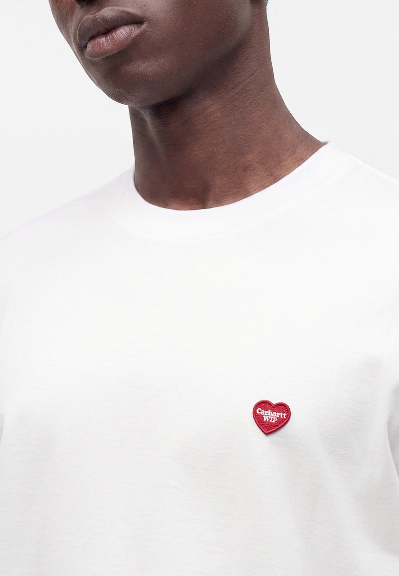Person trägt ein weißes T-Shirt mit einem kleinen roten Herzpatch auf der Brust, auf dem "Carhartt WIP" steht.