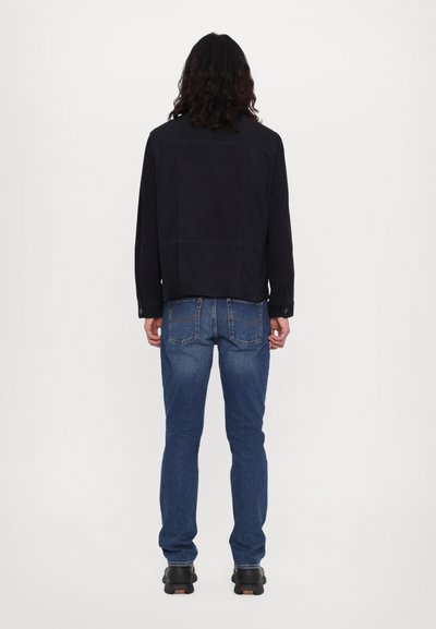 Veste en daim noir avec une coupe boxy, associée à un jean bleu. La veste possède des manches longues et des accents en boutons, tandis que le jean a des poches arrière avec des coutures.