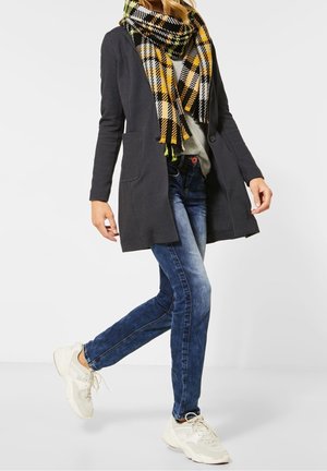 Person wearing a black coat, yellow-black checkered scarf, gray sweater, blue jeans, and white sneakers walking against a plain background.