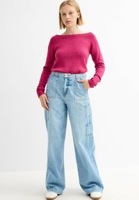 Suéter de punto fucsia con un escote amplio, combinado con jeans cargo azul claro que cuentan con múltiples bolsillos de parche y un corte holgado.