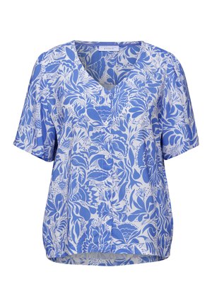 Kvinde iført en blå og hvid blomstret kortærmet bluse og lyseblå jeans, set bagfra med et lille smil.