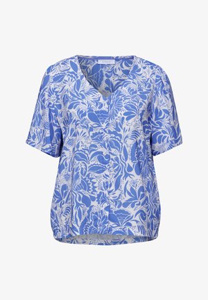 Frau trägt eine blau-weiße Bluse mit kurzen Ärmeln und Blumenmuster sowie helle blaue Jeans, steht mit leichtem Lächeln mit dem Rücken zur Kamera.