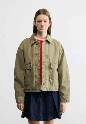 Junge Frau mit glatten braunen Haaren trägt eine olivgrüne, geknöpfte Jacke, ein rotes Oberteil und einen dunklen, plissierten Rock, sie steht vor einem weißen Hintergrund.