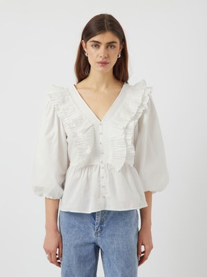 Hvit bluse med volanger, V-hals og knappelukking. Har blafrende ermer og innsamlet midje. Båret med blå jeans.