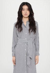 Abito a camicia con righe verticali blu navy e bianche, maniche lunghe, colletto, tasche frontali e un punto vita accentuato con fiocco. Tessuto morbido.