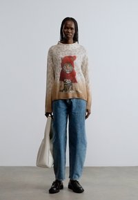 Ülema mõõduga beež kampsun leopardimustriga, millel on värviline joonistus punases mütsis kassist. Sobitatud siniste teksade ja mustade kingadega.