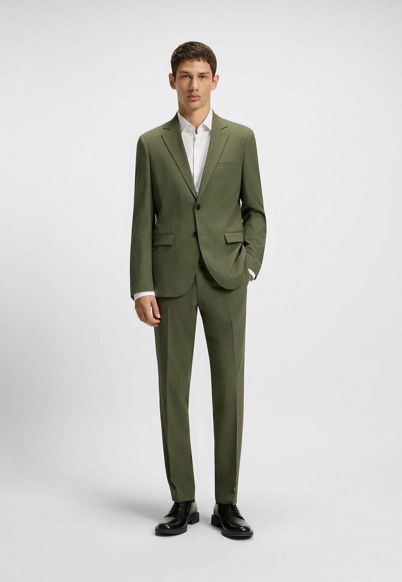 Olivgrüner Anzug mit einreihigem Blazer, aufgesetzten Taschen und schmal geschnittenen Hosen. Kombiniert mit einem weißen Hemd und schwarzen Schuhen.