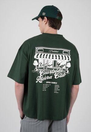 Mann trägt dunkelgrüne Kappe und passendes T-Shirt mit weißer Grafik eines Cafés und Kaffeemenü auf dem Rücken, steht vor einem einfarbigen Hintergrund.