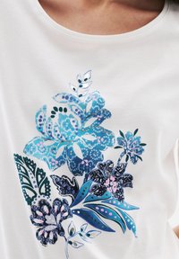Witte t-shirt met een blauwe bloemenafbeelding, inclusief grote bloemen en bladeren met verschillende patronen en schakeringen van blauw, roze en groen.