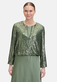 Grüne Pailletten-Bomberjacke mit einem Reißverschluss vorne, ausgestattet mit einem verkürzten Design und langen Ärmeln, getragen über einem passenden grünen Kleid.
