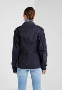 Chaqueta de algodón azul marino con correas en los hombros, puños abotonados y un corte relajado, combinada con jeans azul claro.