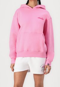 Personne portant un sweat à capuche rose vif avec une poche frontale et un short blanc avec un petit logo coloré sur la jambe gauche.