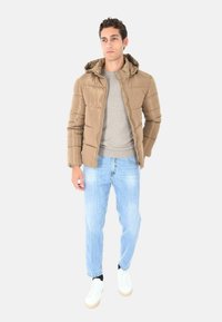 Giacca imbottita marrone con cappuccio, chiusura con zip e polsini a coste, abbinata a un maglione grigio, jeans azzurri chiari e sneaker bianche.