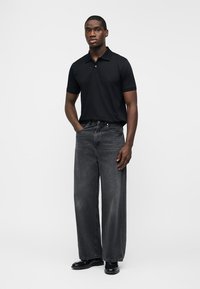 Άνδρας στέκεται φορώντας μαύρο πόλο πουκάμισο, φαρδύ ξεθωριασμένο μαύρο τζιν και μαύρα παπούτσια, μπροστά σε απλό λευκό φόντο.