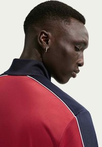 Αθλητική μπλούζα κόκκινου και ναυτικού μπλε χρώματος με γιακά, κατασκευασμένη από υφασμάτινη υφή, με λευκές λεπτομέρειες κατά μήκος των ραφών στους ώμους και εφαρμοστή σχεδίαση.