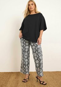 Svart oversized topp i kombination med löst sittande byxor med ormskinnsmönster i svart och vitt; modellen bär svarta sandaletter med remmar på en strukturerad yta.