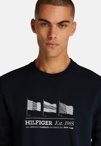 Sudadera azul marino con tres gráficos de banderas blancas, texto "HILFIGER Est. 1985" y un cuello y puños acanalados para mayor textura.