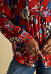 Rote Bluse mit Blumenmuster, langen Ärmeln und gerafften Manschetten, verziert mit blauen und rosa Blumen. Kombiniert mit Jeans und geschichteten Ringen.