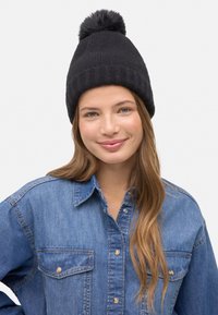 Gorro de punto negro con acabado texturizado y un pompón en la parte superior, llevado sobre una camisa de mezclilla azul claro con botones visibles.