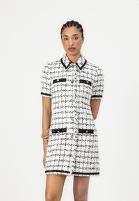 Short-sleeve dress in white tweed with black grid pattern, featuring black trim, button closures, and front pockets with decorative buttons.