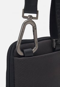 Sort tekstureret taske med lynlås, prydet med en grå metal karabinhage fastgjort til et læderrem. Logo præget på remmen.