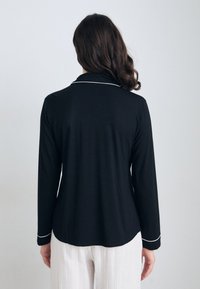 Chemise noire à manches longues avec des garnitures blanches le long du col et des manches. Tissu doux, coupe décontractée, ourlet arrondi, et design à l'arrière visible.
