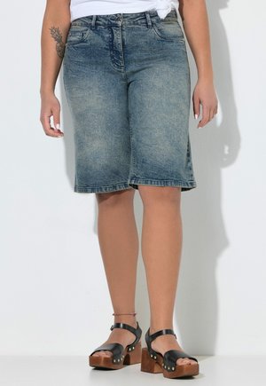 Kvinde iført falmede knælange denimshorts, en hvid knyttet top og sorte platformsandaler med træhæle.