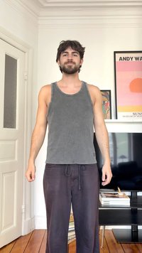 Mann med mørkt hår og skjegg står innendørs iført en grå singlet og løse, mørke bukser i et rom med innrammet kunst og en TV.