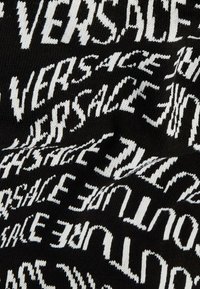 Pleteni materijal sa ponavljajućim tekstom "VERSACE" u bijeloj boji na crnoj pozadini. Tekstura izgleda meko s tijesnim, rastezljivim dizajnom.