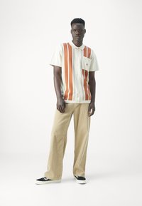 Camisa de botones de manga corta con rayas verticales blancas y naranjas, combinada con pantalones anchos beiges y zapatillas negras. Detalles de tejido texturizado.