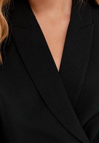 Black blazer featuring a notched collar, structured shoulders, and smooth fabric, showcasing a tailored fit and minimalist design.