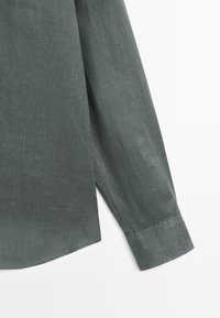 Poignet à manches longues et fente latérale d'une chemise en lin vert foncé sur fond blanc, montrant la texture du tissu et les détails de couture.