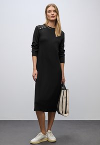 Schwarzes Strickkleid mit langen Ärmeln, Rundhalsausschnitt und Knopfakzenten an den Schultern. Kombiniert mit einer gestreiften Tragetasche und hellen Sneakers.