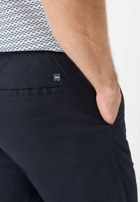 Pantaloni corti neri realizzati in tessuto leggero, con una tasca laterale e un'etichetta con logo discreto. La trama sembra liscia e informale.