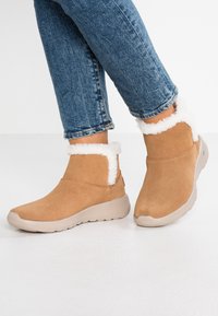 Stivaletti alla caviglia in suede color sabbia con un morbido polsino in ecopelliccia bianca, punta arrotondata e suola flessibile beige, abbinati a jeans blu con orlo arrotolato.