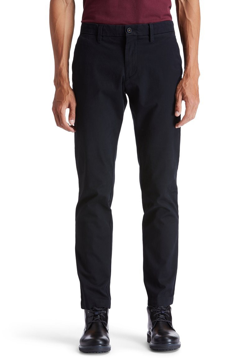 Black cotton trousers with a straight leg, featuring a front button closure, belt loops, and a smooth texture. Worn with black leather boots.