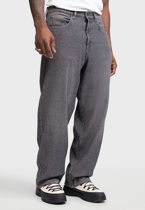 Homme portant un pantalon gris à fines rayures ample avec des chaussures à lacets blanches et noires, debout devant un fond uni.
