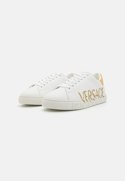 White leather sneakers with gold accents, featuring a logo on the side and a textured rubber sole with a geometric pattern.