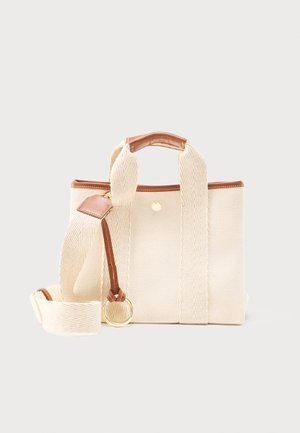 Sac cabas en tissu beige avec une large bandoulière, des garnitures en cuir marron, un détail en anneau doré et une petite fermeture à bouton-pression doré.