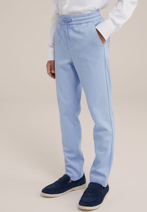 Person iført lyseblå joggingbukser med snor, hvid langærmet skjorte og navyblå slip-on sko står med den ene hånd i lommen.