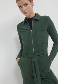 Combinaison en velours côtelé vert avec fermeture éclair à l'avant, deux poches poitrine et une taille ceinturée. Équipée d'un col et d'un tissu texturé.