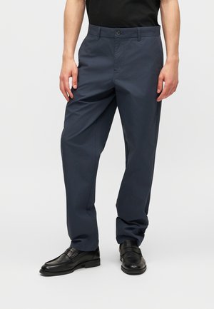 Άνδρας που φοράει ναυτικό μπλε παντελόνι ίσιου ποδιού και μαύρα δερμάτινα μοκασίνια, με απλό ανοιχτό φόντο.