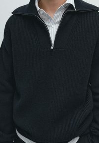 Mörk stickad tröja med kvartslång dragkedja, med ett strukturerat mönster och en lager-på-lager vit randig skjorta under.