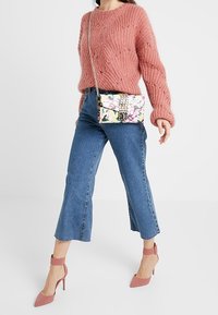 Lyserød strikket sweater, blå cropped jeans, lyserøde højhælede sko og en blomsterpræget taske med guldhardware. Teksturerede materialer og livlige farver.